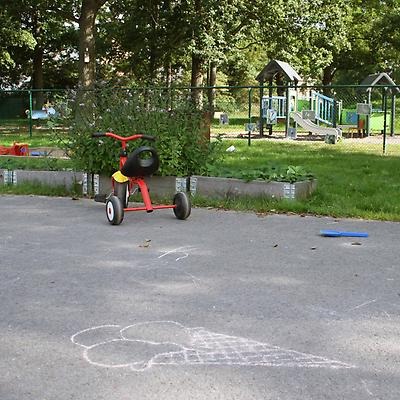 Röd trehjuling vid en lekplats med klätterställning och en glass ritad på asfalten med krita.
